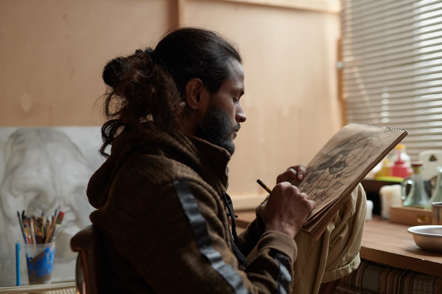 Un artista dibuja en un cuaderno en una mesa de estudio iluminada por el sol, rodeado de pinceles y pintura