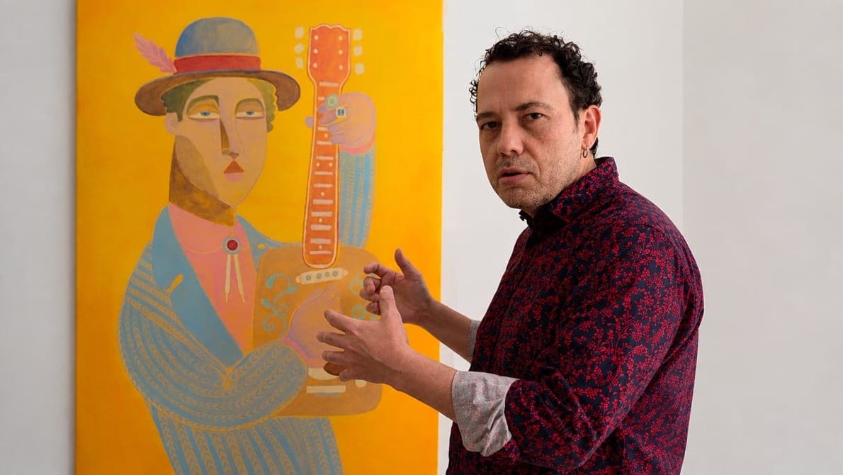 Felix Zilinskas junto a un lienzo amarillo vertical con una figura de sombrero y traje azul que sostiene una guitarra naranja construida como una caja de cerillas, con las manos levantadas en plena explicación