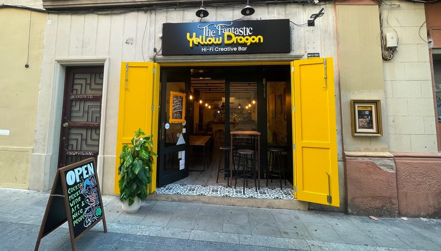 Vista desde la calle de The Fantastic Yellow Dragon, el bar creativo hi-fi de Gràcia, con los postigos amarillos abiertos de par en par y una pizarra en la acera que anuncia Cocktails, Mocktails, Wine, Art & Nice people