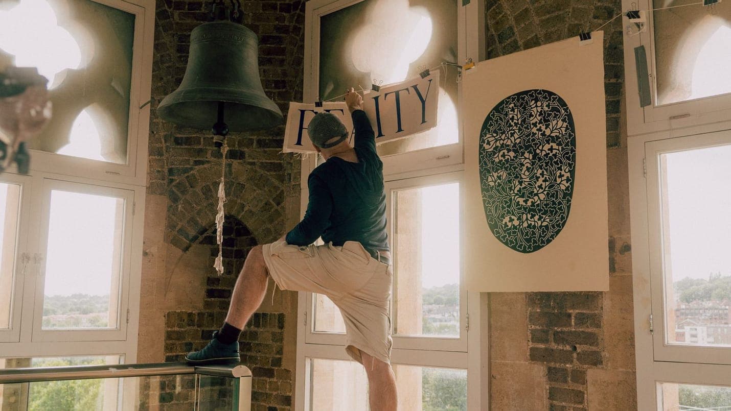 Delehag haciendo equilibrio sobre una barandilla en su estudio de la torre, estirándose para sujetar con pinzas una pancarta con la palabra REALITY escrita a mano junto a una campana de bronce colgante