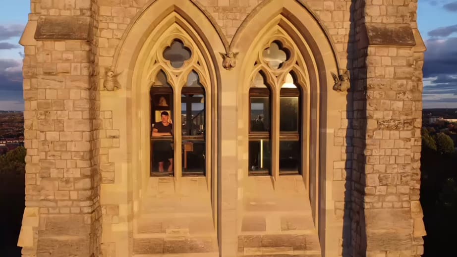 La fachada de la torre a la luz del atardecer: dos altos arcos góticos con tracería de cuadrifolio y ménsulas talladas con ángeles, y al fondo Delehag pequeño en la ventana iluminada